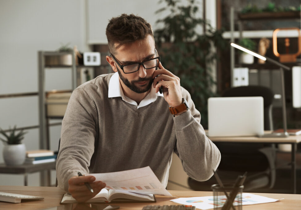 Man talking on phone while working with documents at table in of Man talking on phone while working with documents at table in office